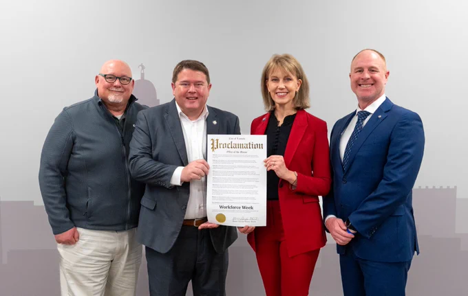Four individuals smiling and standing in a row indoors, with one holding a proclamation document titled "Workforce Week". They are dressed in business attire.