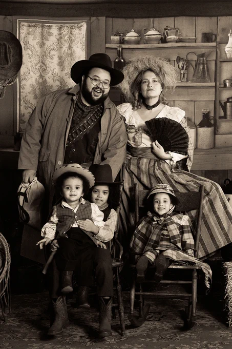 From Severance. A sepia photo of Dylan (portrayed by Zach Cherry) and his wife Gretchen (portrayed by Merritt Wever) posing for the camera while wearing clothes that look like they're from the Wild West era in America. They appear happy, and their three kids are sitting below them smiling.
