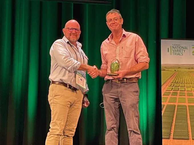 Andrew Russell (left) and Cam Nicholson (right) shake hands in front of a dark green curtain on a stage. Cam is holding an award. 