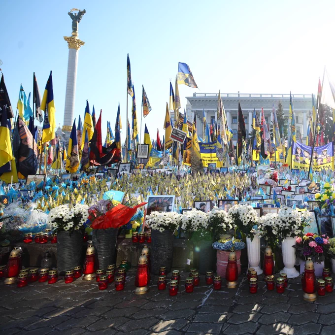 A memorial site filled with flowers, candles, and numerous Ukrainian flags stands in at Maidan square in Kyiv. Photographs of individuals, messages, and banners are displayed in tribute to those honored. In the background, the Independence Monument and a neoclassical building are visible. The scene is illuminated by sunlight, emphasizing the solemn yet powerful atmosphere of remembrance. 
