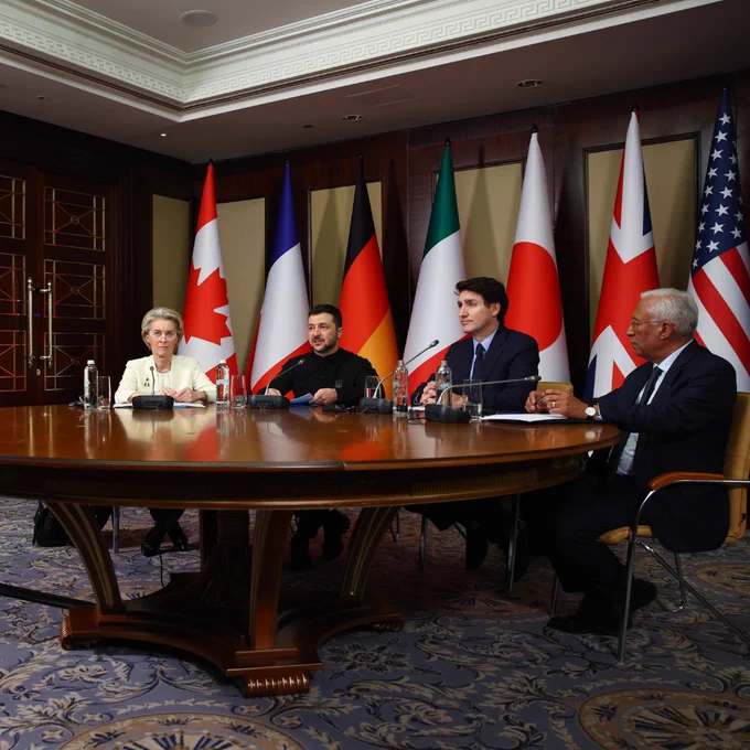 From left to right, President von der Leyen, President of Ukraine Volodymyr Zelenskyy, Canadian Prime Minister Justin Trudeau and EU Council President Antonio Costa sit at a round wooden table during a formal meeting, with microphones and water bottles in front of them. Behind them, flags from Canada, France, Germany, Italy, Japan, the United Kingdom, and the United States are displayed.