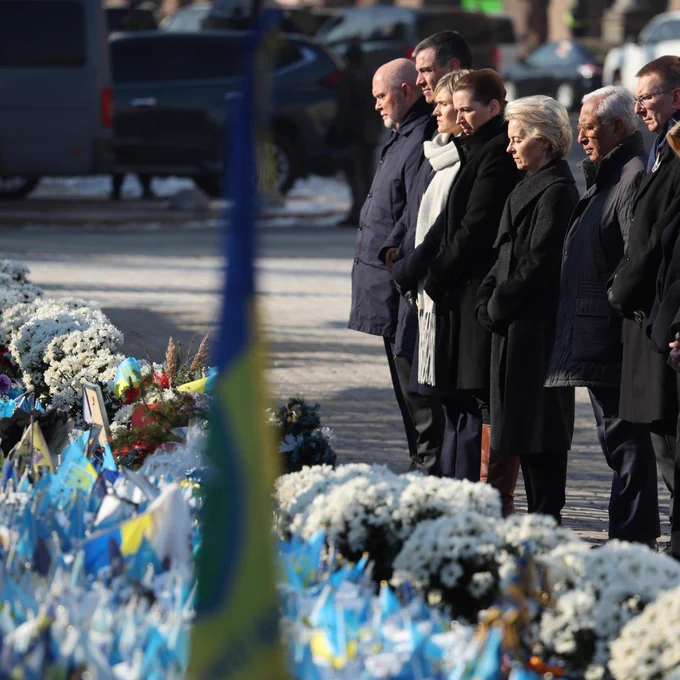 President von der Leyen together with President Antonio Costa and officials stand in a solemn tribute before a memorial adorned with white flowers and blue-and-yellow flags. They are dressed in dark winter clothing, with serious expressions, as they pay their respects. 
