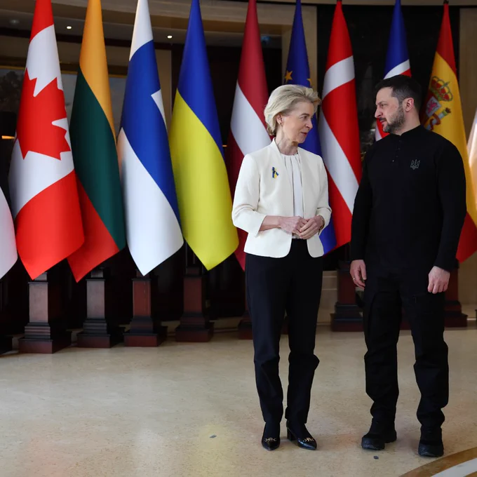 President von der Leyen talking with President of Ukraine Volodymyr Zelenskyy. Behind them, flags from Canada, Lithuania, Finland, Ukraine, Latvia, Europe, Denmark, UK and Spain are displayed. 