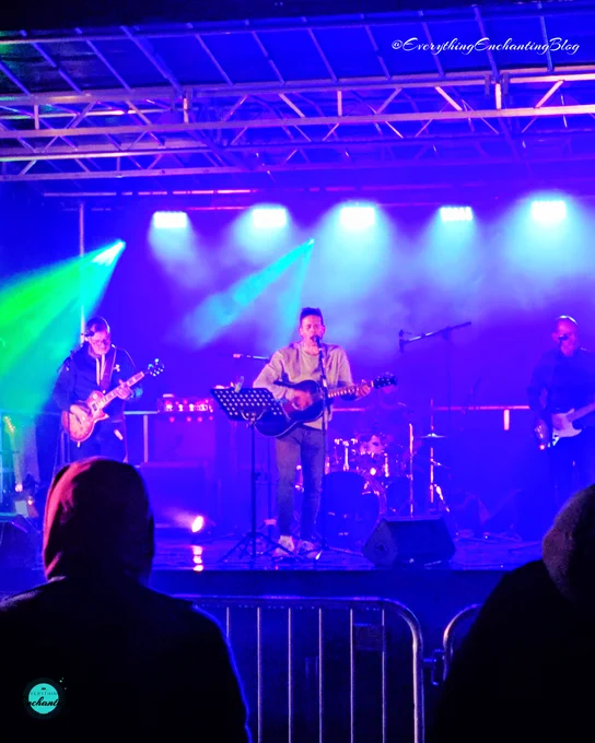 The Tom Betts Band is performing on a stage with vibrant blue and pink lighting. The band members are playing their instruments, and the lead singer is at the center with a microphone stand. The stage is framed by a metal structure, and there is a crowd of silhouetted audience members in the foreground. The lighting and fog effects add to the energetic ambiance of the live performance