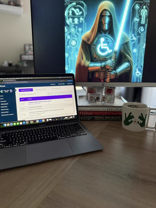 Computer desk with screen open to axe-con, a asl coffee mug to the right of the computer and larger adapted computer screen with an a11y Jedi on it