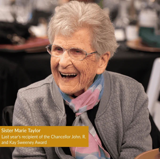 Sister Marie Taylor is the last year's recipient of the Chancellor John. R. and Kay Sweeney Award. She is seated at a table, engaged in conversation. The background suggests a social gathering with blurred tables and chairs.