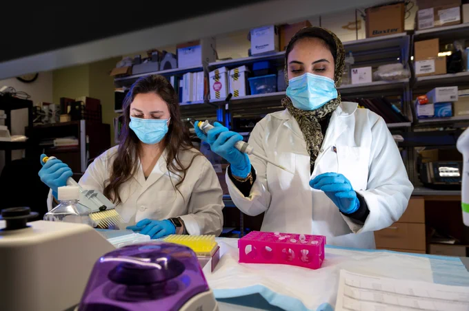 Two women in lab coats and surgical masks stand at a lab bench using pipettes and other lab equipment.