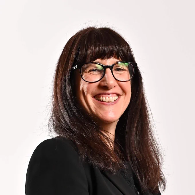 An image of a woman smiling. She has dark brown hair past the shoulder and glasses.