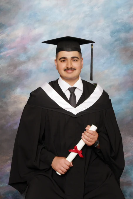 A man stands posed wearing grad regalia, while holding a certificate in his hands.