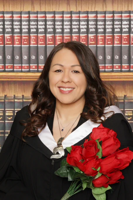 A woman dressed in grad regalia holds roses and stands in front of book shelves.