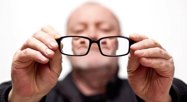A man looks through his glasses while holding them away from his face.
