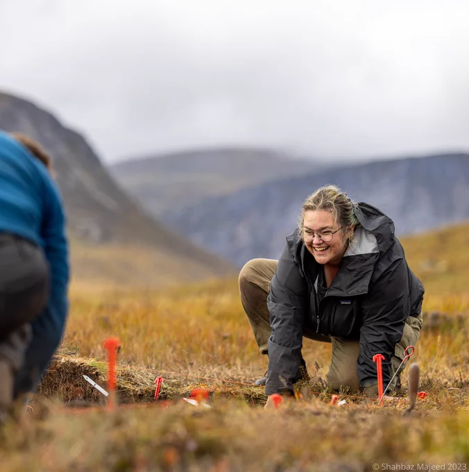 Smiling person leaning down by a trench in a mountain setting