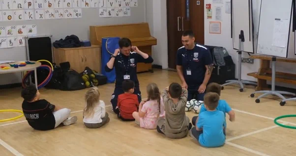 Children in a sports hall taking part in a literacy activity with community football club staff leading 