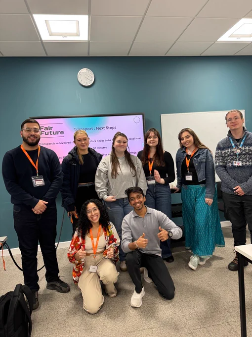 A group of 8 student officers smiling at the camera. They are in a room with teal walls, stood in front of a presentation thats titled "Transport: next steps."