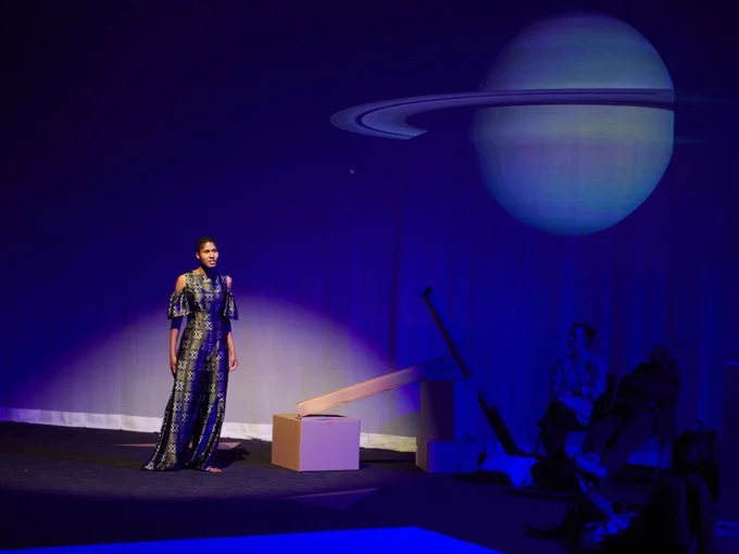 Professor Danielle Wood stands in the spotlight on a darkened stage. An image of Saturn is projected on the wall to her left. Credit: Doug Segars