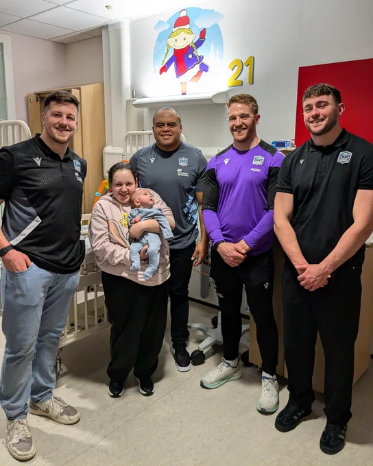 4 Glasgow Warriors rugby players stand behind a mum holding her baby in a hospital room. the baby is staring up at the players, who are all smiling at the camera.