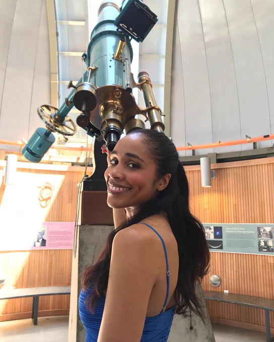 Nia is similar at the camera in a blue dress while the telescope is behind her.