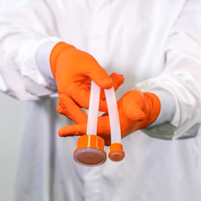 A person in a lab coat and orange gloves holding two test tubes.