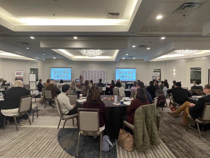 A photo from the back of a hotel meeting room showing many people seated at round tables and two screens at the front of the room displaying slides.