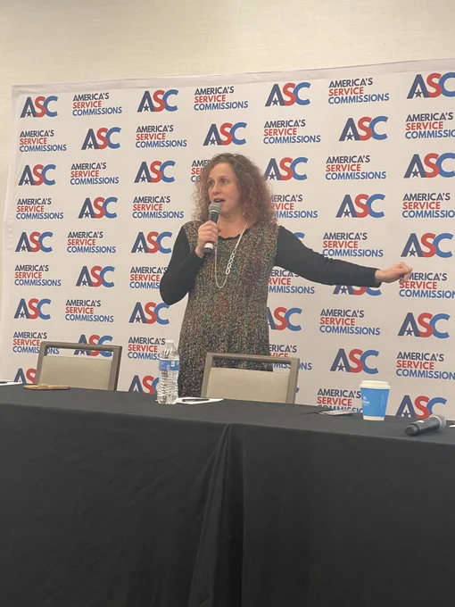 A photo of a person standing behind a long table and in front of an ASC step-and-repeat backdrop. The person is speaking into a microphone.