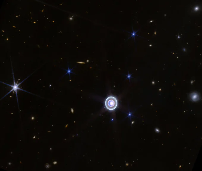 A wide-field image of Uranus (near the center), taken in the infrared by JWST, shows the glow of the perpendicular rings around the planet. Several stars can be seen and recognized by their tell-tale diffraction spikes. 14 small dots are the moons of Uranus. And everything else in the image is a distant galaxy, ranging from on-edge spirals to faint blobs.