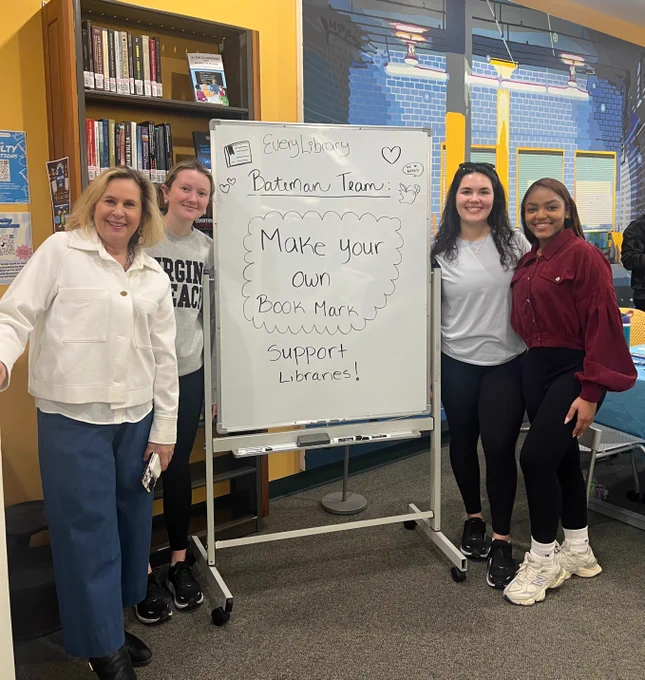 Four individuals posing in front of a whiteboard with text promoting a bookmark creation event to support libraries at the EveryLibrary.