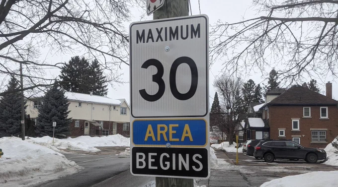  A picture of a speed limit sign attached to a pole with a snowy background and some houses and parked cars.  The Sign Reads "Maximum 30" in black on white, then "Area"  in yellow on blue, then "Begins" in white on black. photo: dan kellar
