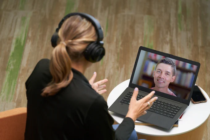 Person attends a virtual meeting with Ryan Seacrest displayed on the laptop screen, while sitting at a wooden table and wearing headphones.