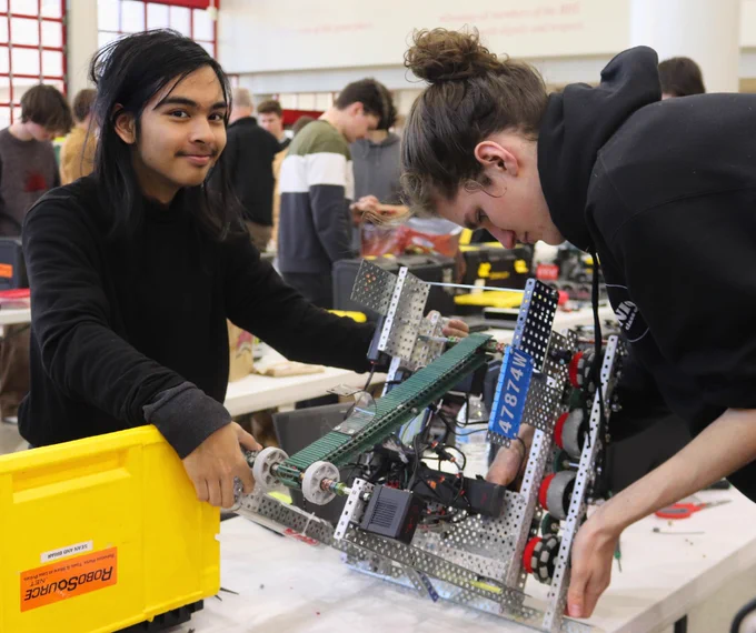 A student looks toward camera while helping another student work on a robot on a table