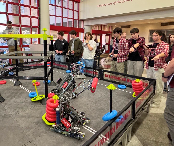 Group of students stand around robotics stage field and control robots in action