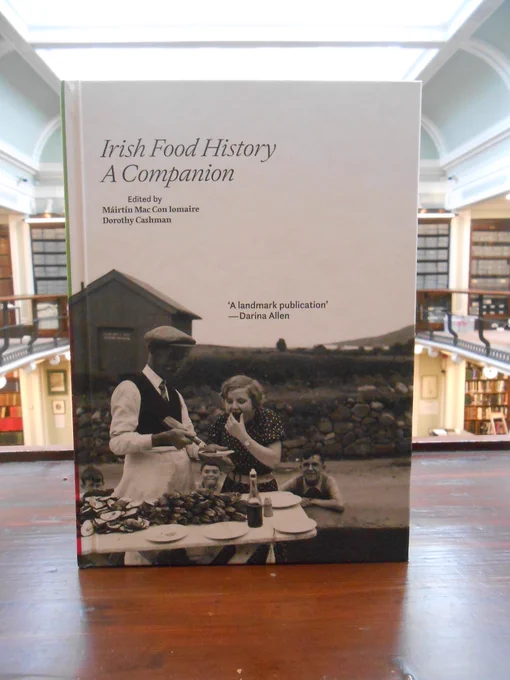 A book entitled 'Irish Food History: A Companion' edited by Máirtín Mac Con Iomaire and Dorothy Cashman. The front cover is a black and white photograph of a man serving a woman oysters on a country road with three young boys looking on.