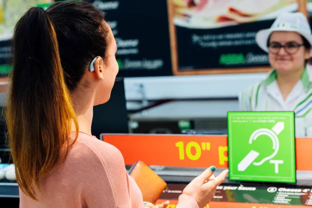 A young woman with a ponytail seen from behind as she stands at the pizza counter in a supermarket. She has a hearing aid and is talking to the staff member behind the counter. There is a hearing loop sign on the top of the counter in the green of the supermarket's brand.