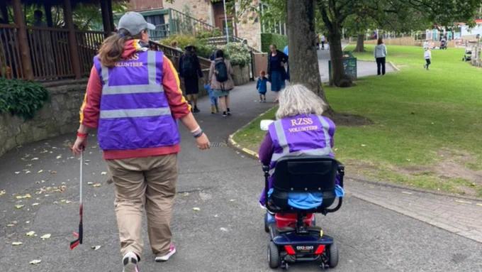Two volunteers make their way around Edinburgh Zoo, one walking and one in a motorised chair. They are wearing purple vests that say "RZSS volunteer".