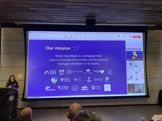 Teams call projected on to a large screen. Shared slide reads “Our mission. Make Your Mark is a campaign that aims to increase the number and diversity of heritage volunteers in Scotland.” Group logos, including Glasgow Disability Alliance. Participants on Teams visible on screen. Rosie Wylie is standing at the lectern, speaking.