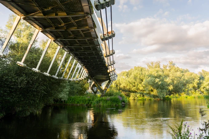 Photograph taken under a long pedestrian bridge spanning a wide river lined with dense, mature trees