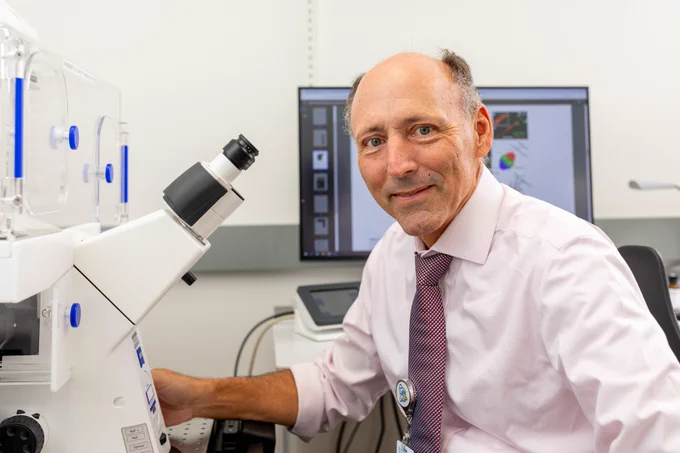 A photo of Dr. Kulesa sitting at a table with a microscope. He is wearing a light pink button-up, collared shirt. There is a computer monitor on behind him.