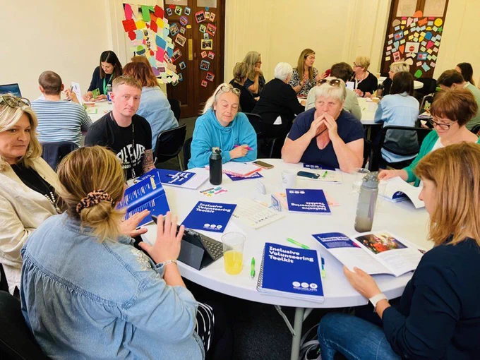 Image depicts a group of people sitting at a white, round table, with bright blue booklets in front of them.  The photo is of a Make Your Mark Inclusive Volunteering Toolkit workshop session.