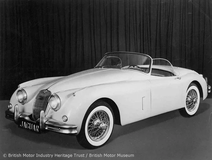 A black and white photograph of a three-quarter view of a white Jaguar XK150 in a studio with a curtain in the background. 