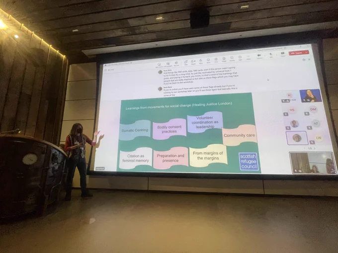 People sat in an auditorium and facing a screen with a presentation from the Scottish Refugee Council.