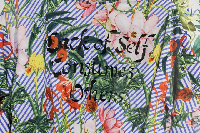 Image: Joiri Minaya, I can wear tropical print now #5 (detail), 2018. Found used shirt, digital print on spandex, custom wooden frame, 32 x 40 x 3 in. Image courtesy of Red Bull House of Art.
 
Description:
 
A close-up of the back of a colorful Hawaiian shirt with light blue and white stripes under pink and orange flowers. Written in black on top: "Lack of Self / Consumes / Others"
 