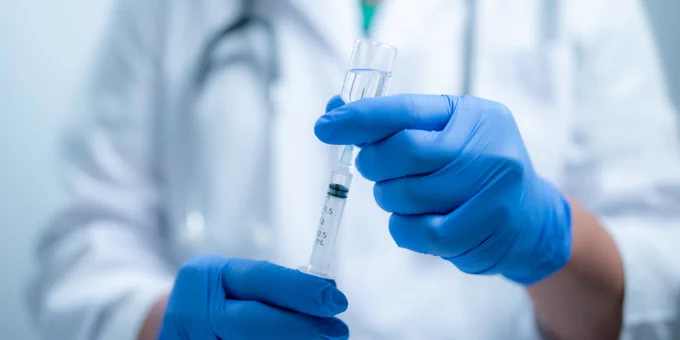A stock image of hands wearing blue gloves holding a needle and vial.