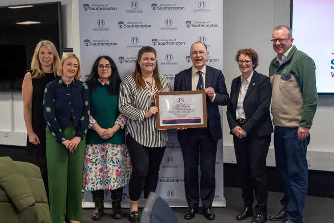 A group of seven people stand together in front of a branded University of Southampton backdrop, posing for a photo. They are dressed in a mix of business and smart-casual attire. The central figure, a woman in a striped blouse, is holding a framed certificate alongside a man in a suit and tie, who is also holding the award. The certificate is labeled "University of Sanctuary Award." The group is smiling, and the setting appears to be an indoor event space with a presentation screen visible on the right side of the image. 