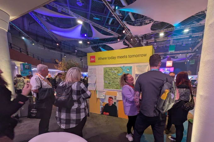 Image of a group of people chatting in a large exhibition hall at NIA in front of the AmplifyFE map.