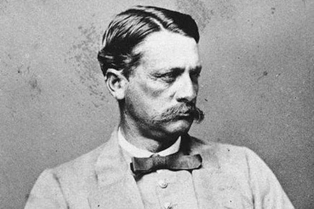 A black and white photo of a man in 1865 with slick back dark hair and a long dark moustache. He is wearing a suit jacket and shirt with a dark coloured bow tie.