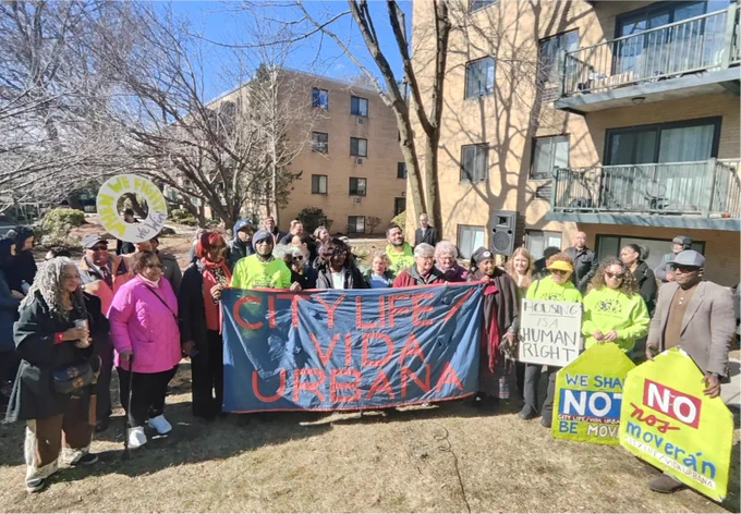 The image shows a large group of people gathered outside in front of a brick apartment building. The group is diverse in age, gender, and ethnicity, and they appear to be participating in a housing rights protest or rally. Many participants are holding signs and banners advocating for housing justice.

At the center, a large blue banner with red text reads:
"CITY LIFE VIDA URBANA"

To the right, a person holds a sign with black text on white that says:
"HOUSING IS A HUMAN RIGHT"

Two additional signs, shaped like houses, read:
"WE SHALL NOT BE MOVED"
"NO nos moverán"
Both of these signs include the name "City Life / Vida Urbana."

Some participants are wearing bright neon green shirts, possibly signifying organization volunteers or leaders. Others are wearing warm clothing, indicating a cold day. Several individuals wear sunglasses, and a few hold walking canes. In the background, apartment balconies and windows are visible.

The mood appears strong and unified, with people standing to