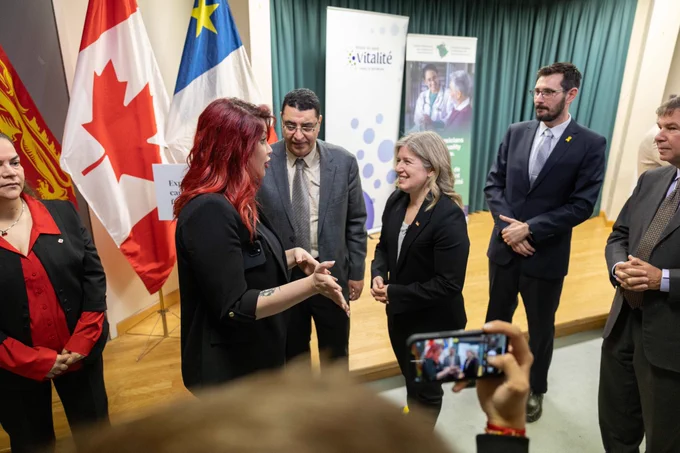 The Premier engaged in conversation with graduates on a stage with Canadian and provincial flags. One person is taking a photo with a smartphone. Banners displaying the word "Vitalité" are in the background. 
-- 
Le première ministre aves les dix nouveaux médecins en tenue de soirée est debout et discute. Des drapeaux canadiens et étrangers sont visibles à l'arrière-plan. Une personne au premier plan prend une photo avec son smartphone. Des banderoles avec du texte et des images sont également visibles. 