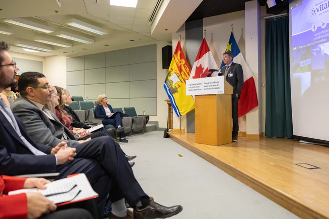 Minister Dornan stands at a podium giving a presentation in a conference room. Flags and a projector screen are behind him. Audience members are seated, listening attentively, some taking notes. 
-- 
Ministre Dornan se tient à une tribune et s'adresse à un auditoire, avec les drapeaux du Nouveau-Brunswick, du Canada, et de l'Acadie derrière lui. L'auditoire est assis attentivement, certains prenant des notes, dans une salle de conférence où des diaporamas sont projetés sur un écran. 