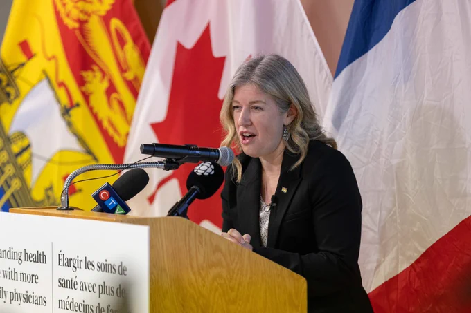 Premier Susan Holt in a black blazer speaks at a podium with microphones. Flags are in the background. The podium has text about expanding healthcare. 
-- 
Première Susan Holt s'exprime sur une tribune équipée de plusieurs microphones, devant des drapeaux canadiens et provinciaux. Elle porte une veste noire et affiche une expression engagée lorsqu'elle s'adresse à son auditoire. 
