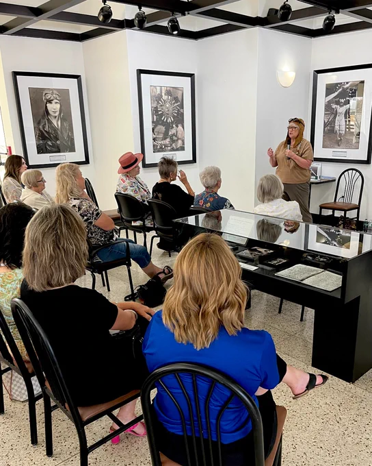 Bird Thomas is giving a presentation to a seated audience in an exhibit dedicated to Vera Dawn Walker with various framed photographs on the walls.