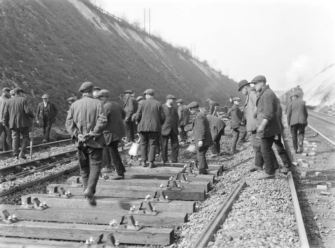 1923 photograph showing gang of track workers relaying a railway line in a deep cutting. A steam train approaches on an adjacent line. Around 20 men are in the scene. Photograph courtesy National Railway Museum.